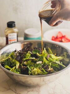 Drizzling vinaigrette over strawberry spring mix salad in large mixing bowl.