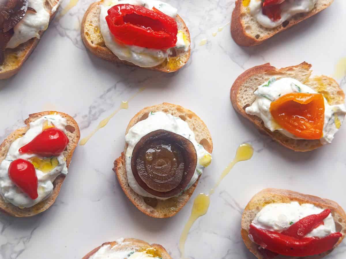 Whipped ricotta crostini with antipasto toppings on white marble table top with drizzled olive oil.