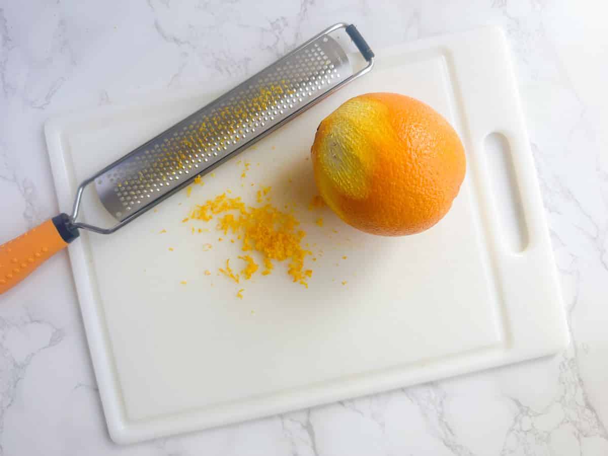 An orange, a microplane, and orange zest on a cutting board.