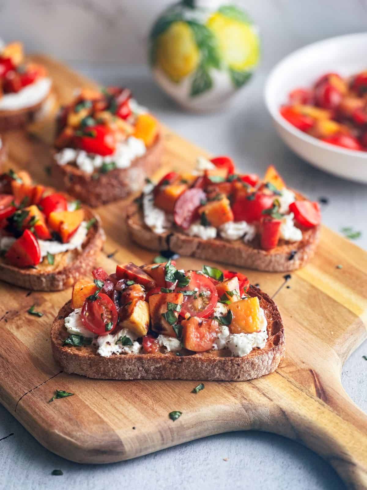 Peach and tomato burrata bruschetta on a wooden board with a bowl of peaches and tomatoes in the background.