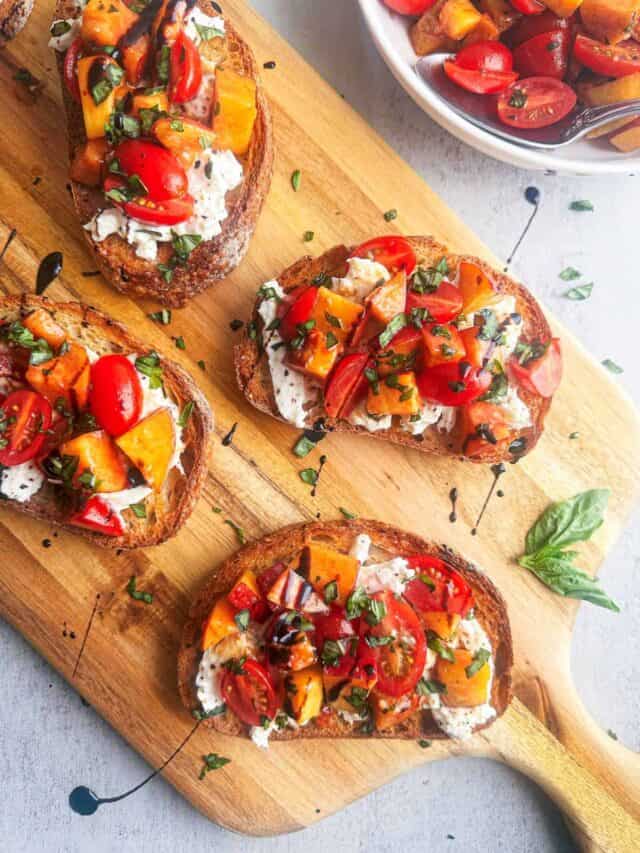 Peach, tomato, basil, and burrata on toasted bread slices on a wooden board.
