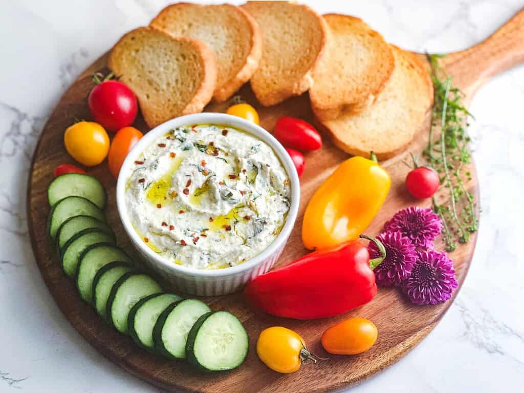 Whipped ricotta dip in a ramekin on a wooden charcuterie board with veggies and crostini.