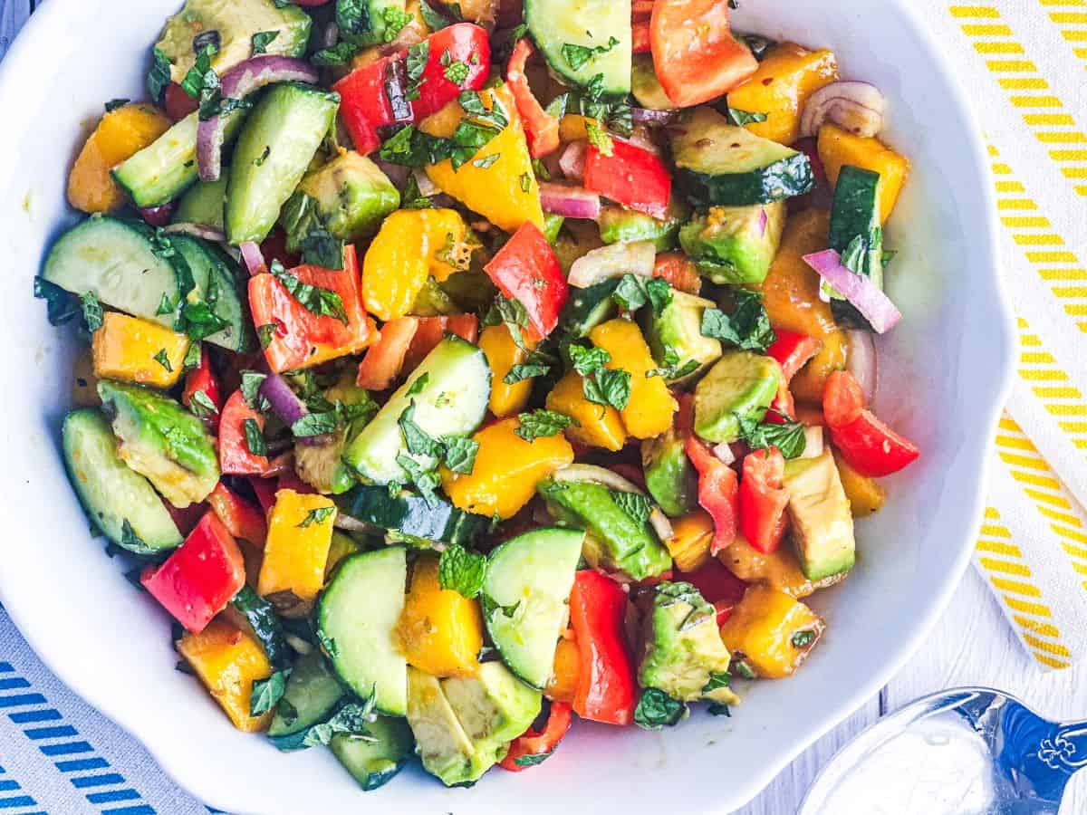 Avocado, cucumber, mango, and bell pepper salad with shallots and fresh mint in a white bowl with a spoon on the side.