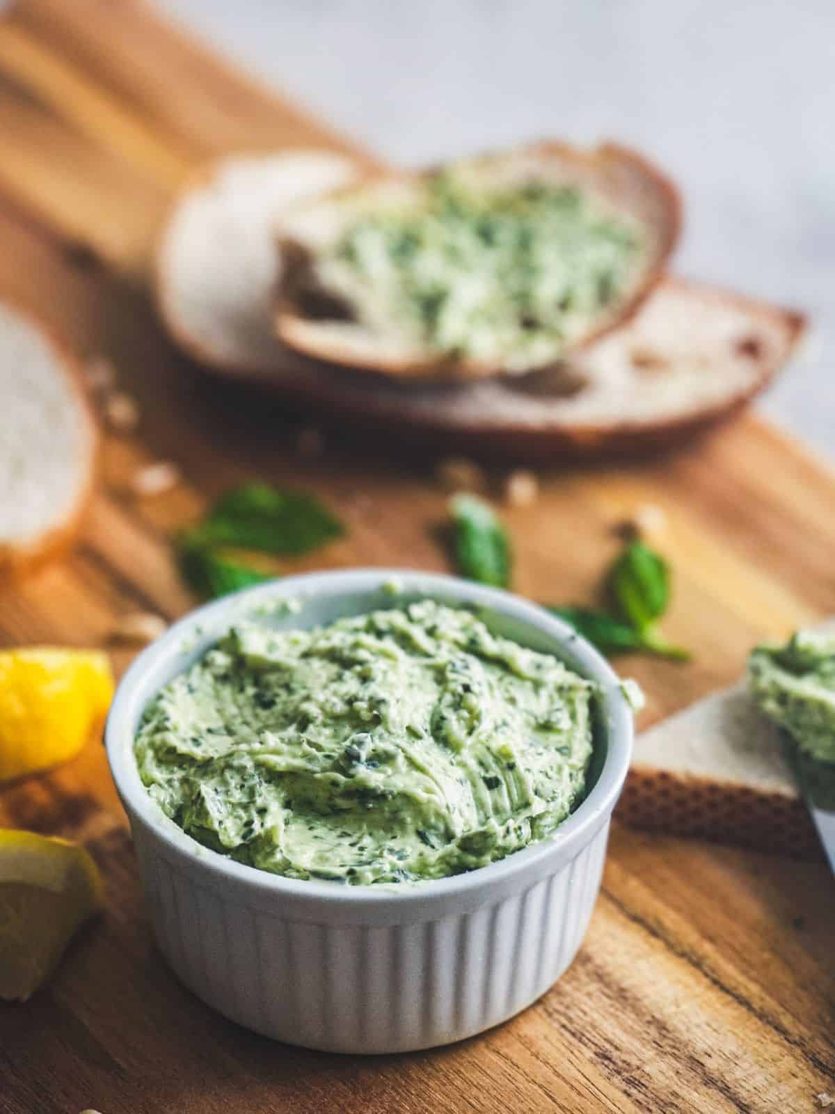 Whipped basil pesto butter in a white ramekin with bread and basil leaves in the background.