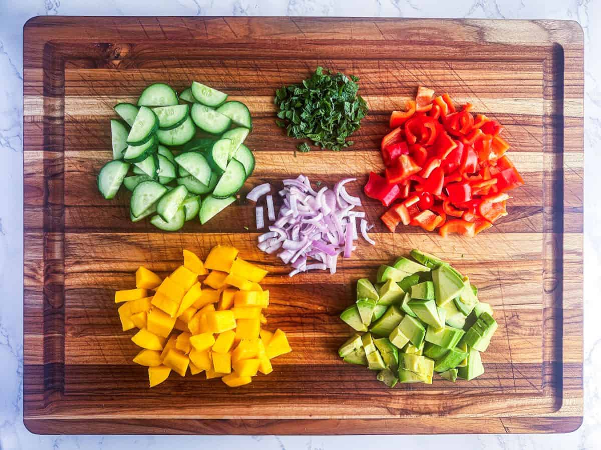 Chopped cucumber, mango, avocado, bell pepper, shallots, and mint on a wood cutting board.