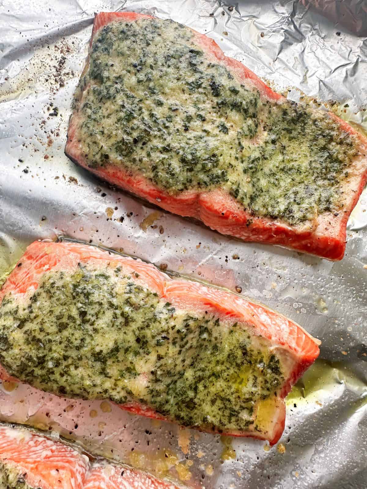 Salmon fillets topped with melted pesto butter on a foil baking sheet.