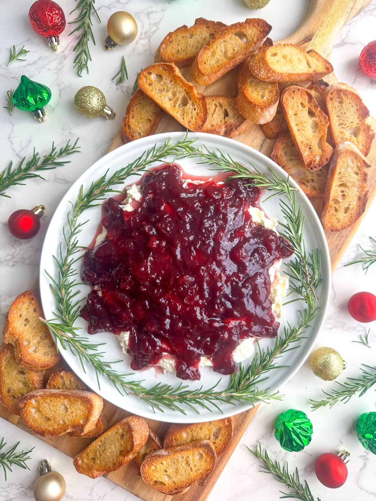 Cranberry feta dip in white bowl with rosemary sprigs, toasted baguette slices, and small Christmas ornaments surrounding them.