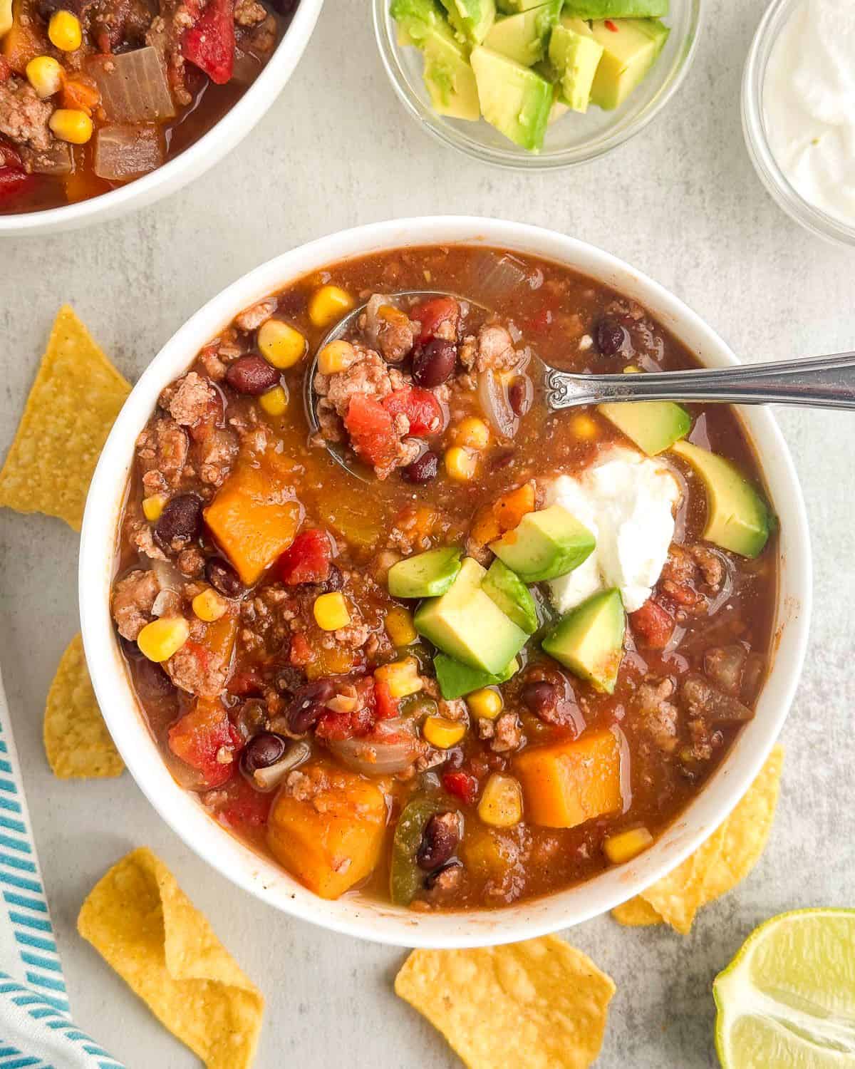 Chili with sweet potato and squash in a white bowl with a spoon, surrounded by tortilla chips.