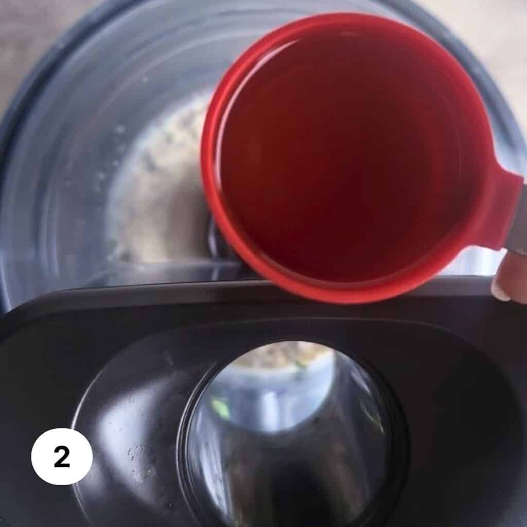 A red measuring cup with oil over the pour-in spout of a food processor bowl.