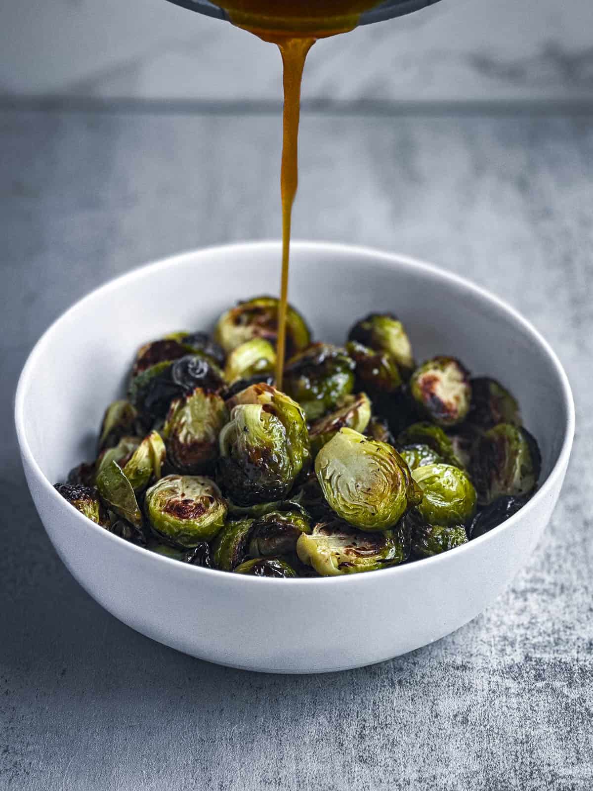 A bowl of roasted brussels sprouts with maple sriracha glaze being drizzled on top.