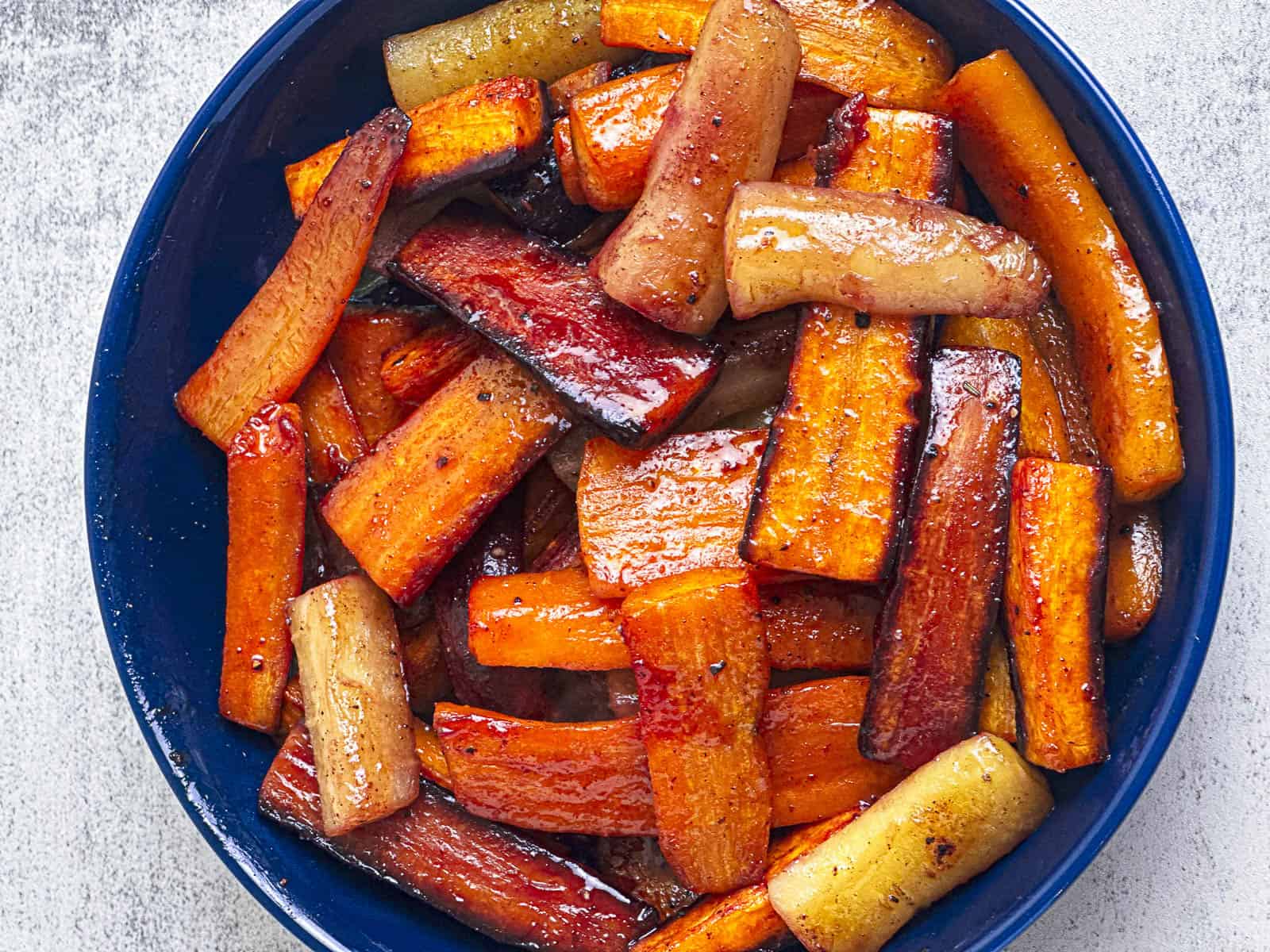 A blue bowl filled with a colorful assortment of roasted root vegetables. The vegetables, including carrots and parsnips, are caramelized with a glossy, browned exterior, indicating they are seasoned and cooked to perfection. The bowl sits on a light gray surface.