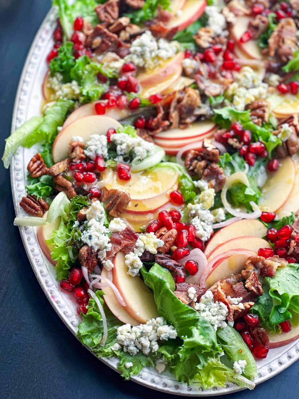 Fresh apple, bacon, pecan, blue cheese, and mixed greens salad with pomegranate arils on a white platter.