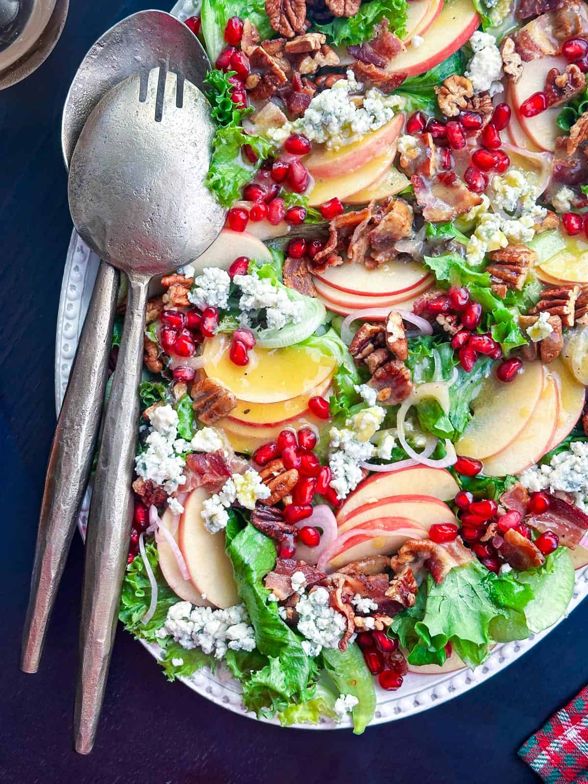 Fall apple salad with pomegranate seeds, blue cheese, pecans, and bacon on a white platter with two serving spoons.