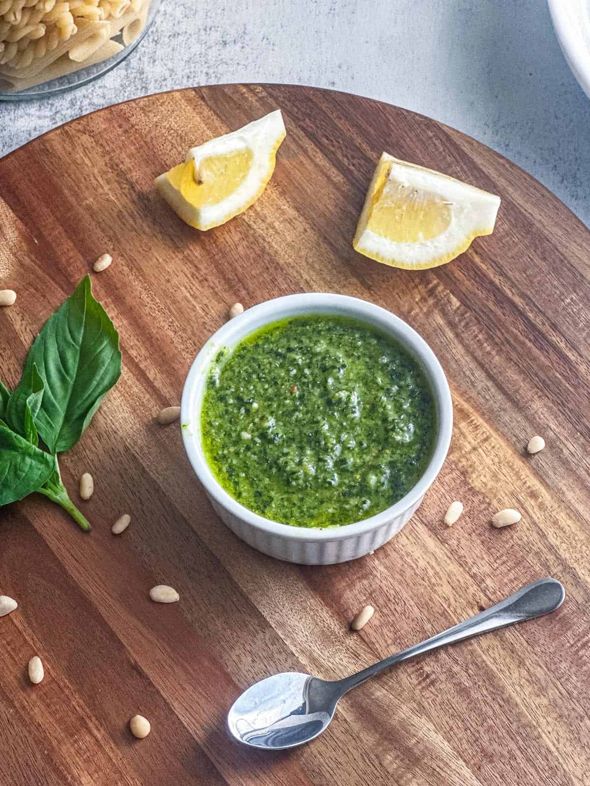 Fresh basil pesto in a ramekin on a wooden board with lemon wedges and pine nuts. Perfect for Italian cuisine or pasta dishes.