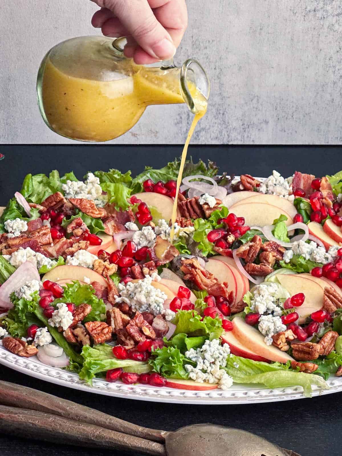 Apple cider vinaigrette dressing being poured over a fresh, colorful Christmas salad featuring pomegranate seeds, pecans, pears, and blue cheese.