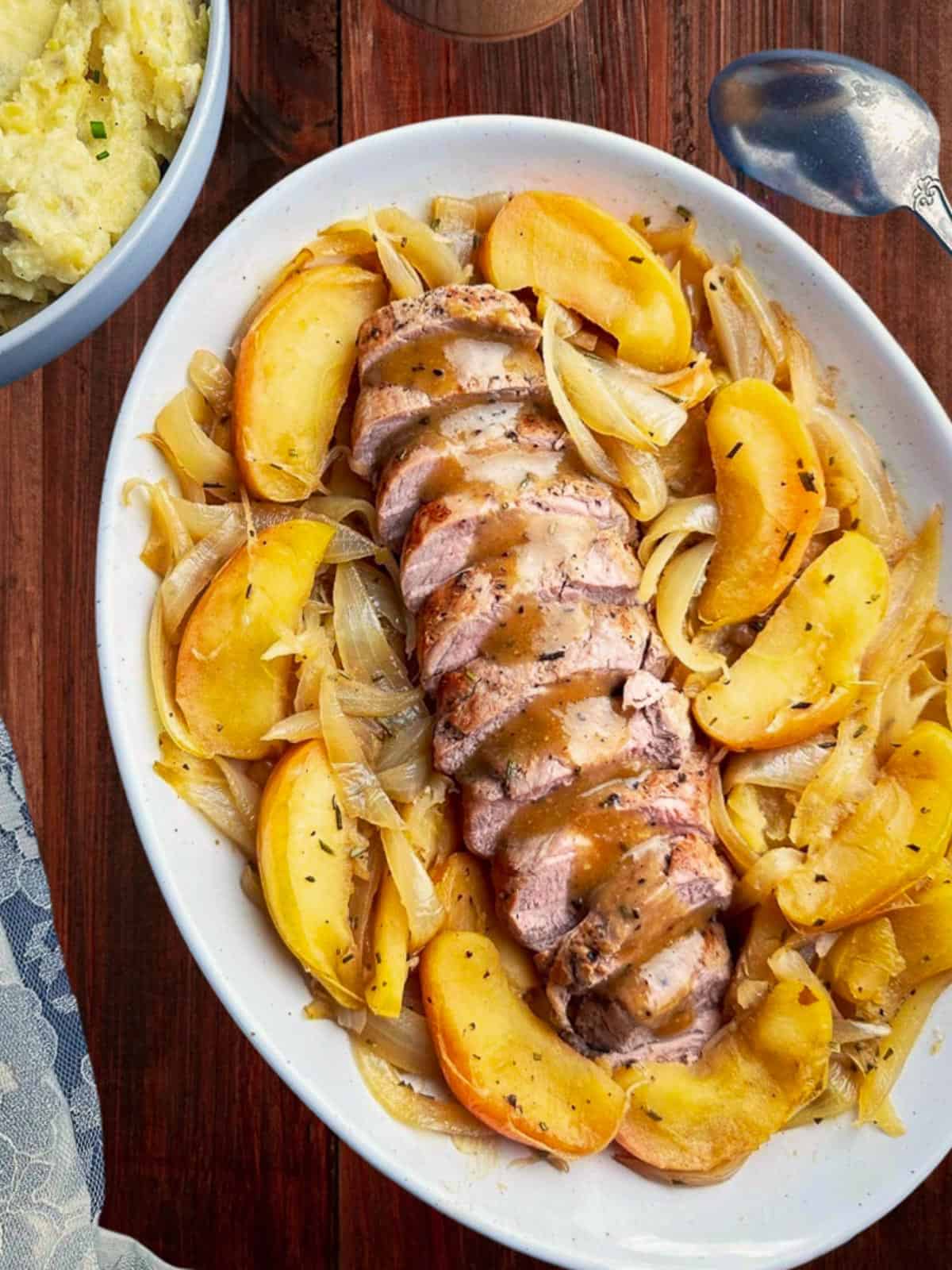 Juicy roasted pork tenderloin slices surrounded by roasted apples and caramelized onions on a white serving dish.