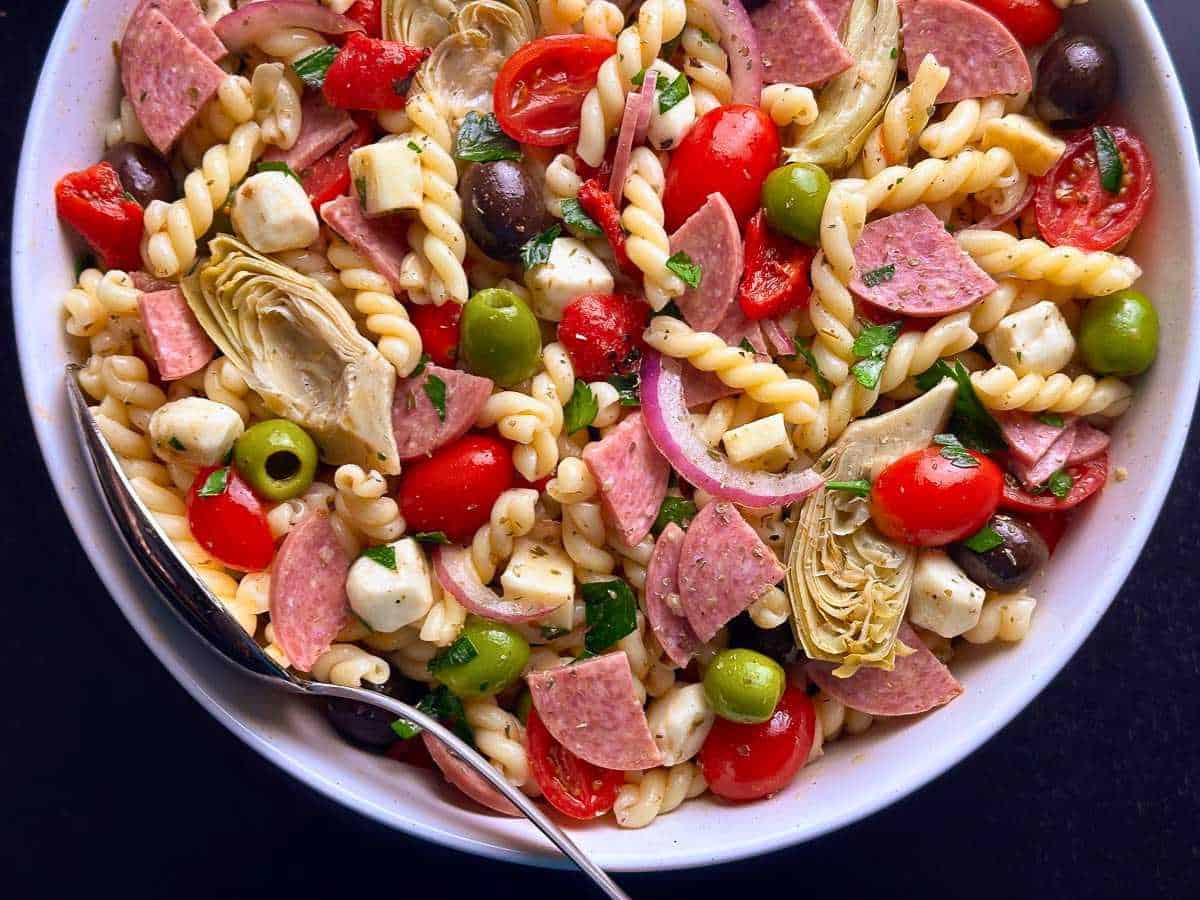 A vibrant pasta salad featuring spiral pasta, cherry tomatoes, olives, artichokes, and salami slices, ideal for picnics and casual dining.