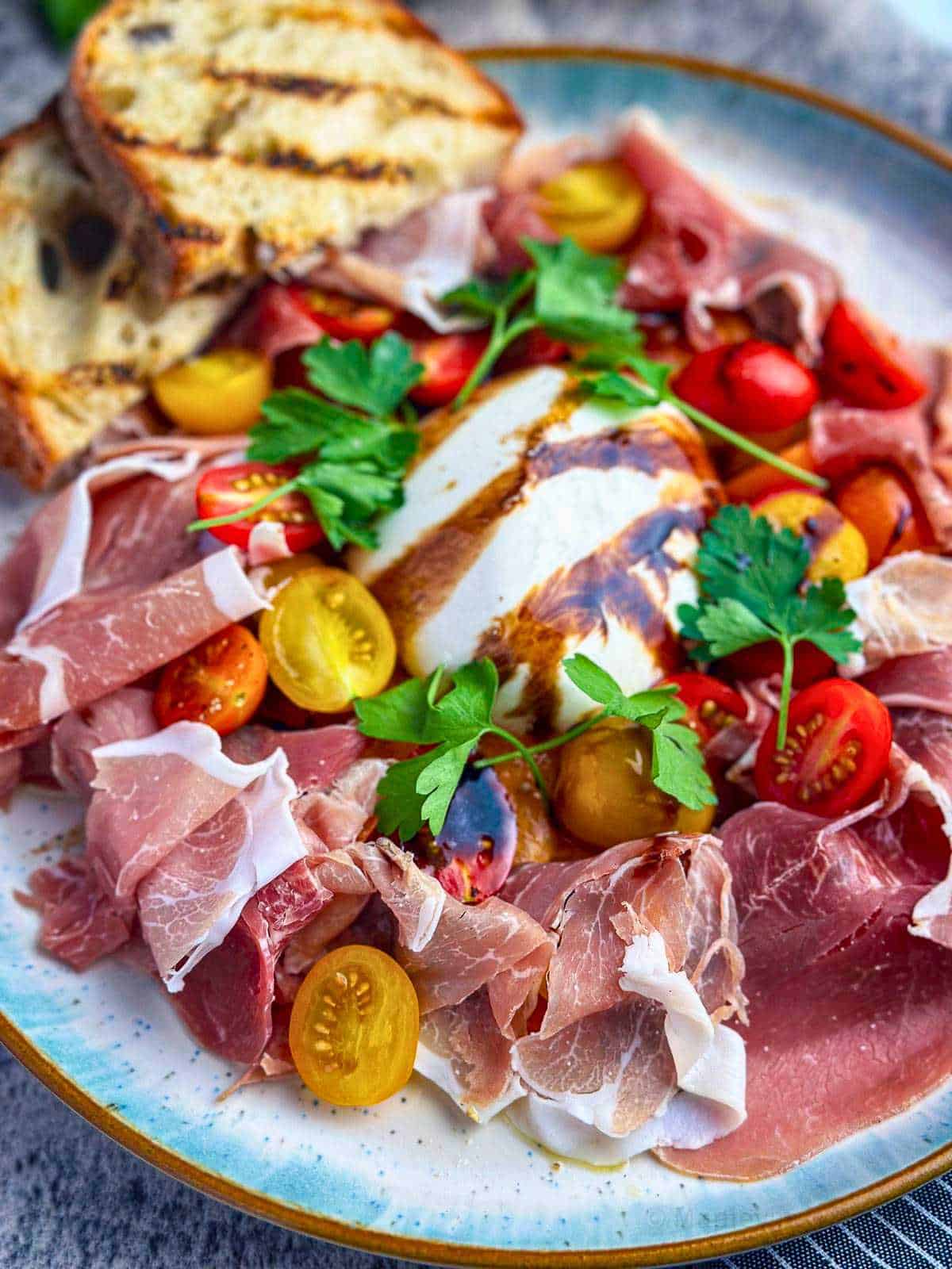 Burrata and prosciutto on a stylish plate with heirloom cherry tomatoes and fresh parsley with a balsamic glaze drizzle.