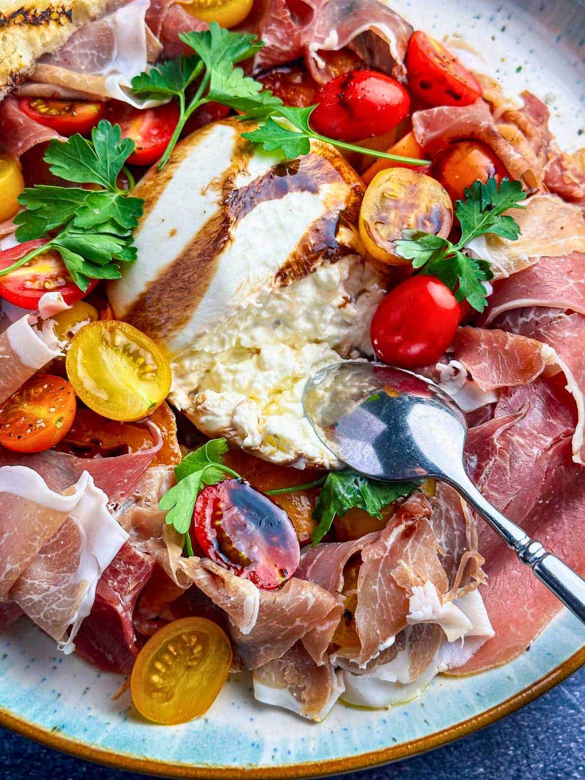 Burrata and prosciutto on an elegant plate with heirloom cherry tomatoes and fresh parsley with a balsamic glaze drizzle.