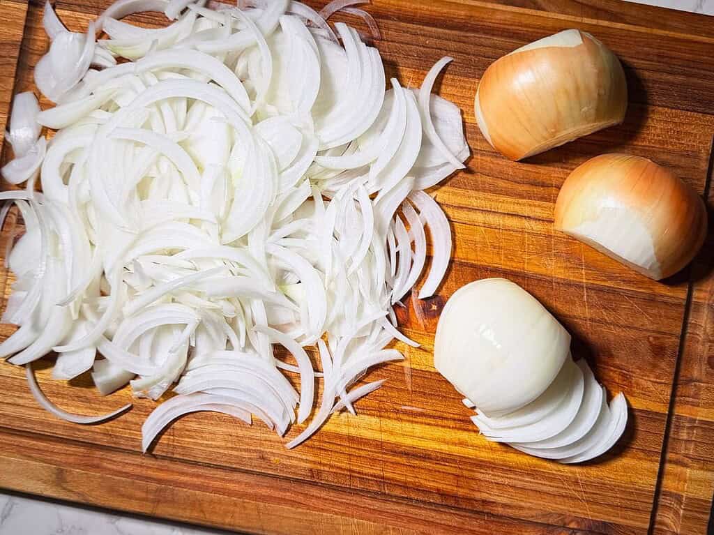 Yellow onions halved and sliced on a wood cutting board.